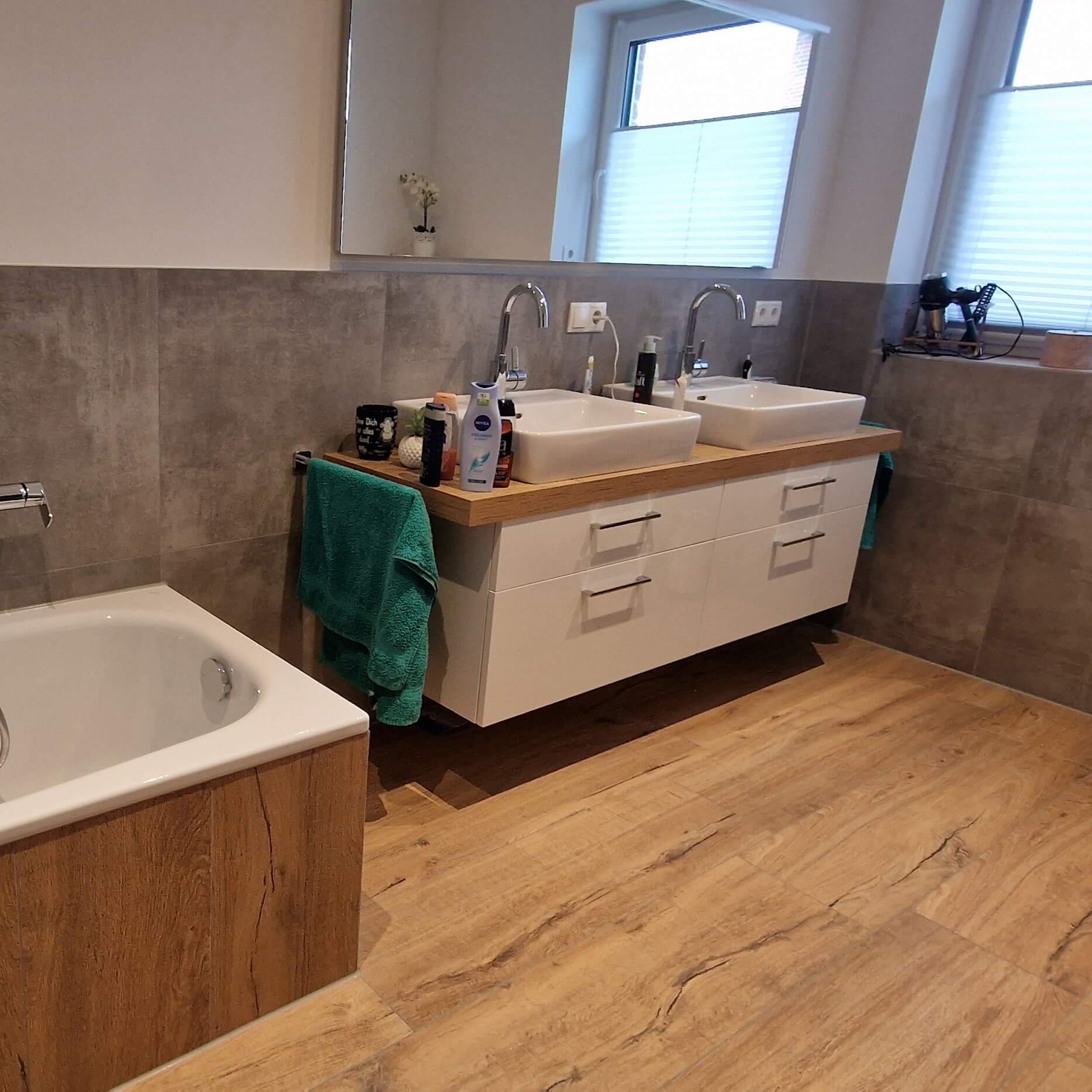 Badezimmer mit Badewanne, grauen Wandfliesen und brauen Bodenfliesen sowie zwei Waschbecken und ein Spiegel.