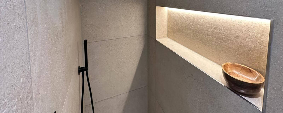 Moderne Dusche mit Sandsteinfliesen und einer in der Wand eingebauten und beleuchteten Ablage auf dem eine Holzschale steht. Gegenüber der Ablage ist ein Brauseset mit Wandanschlussbogen und Brausehalter angebracht.